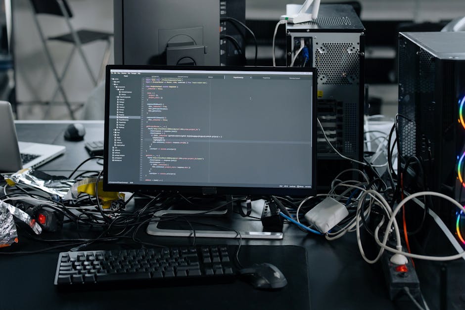 A person seen from behind working at a desk with a laptop displaying lines of colorful code, surrounded by a desktop monitor, a closed folder labeled 'Procurement,' and a black remote control. The wor