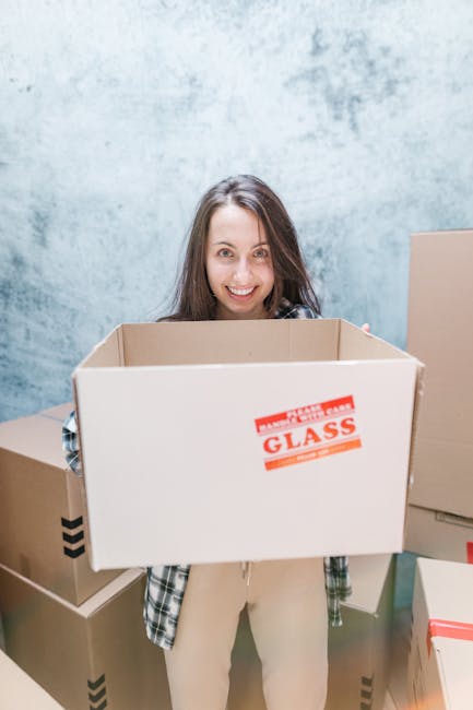 A young woman with long dark hair sitting cross-legged on a wooden floor inside a room filled with packed cardboard boxes and packing materials, including white foam packing peanuts. She is wearing a 